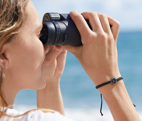 The 4ocean Osborne Reef Bracelet is worn by a female model at the beach as she uses binoculars.